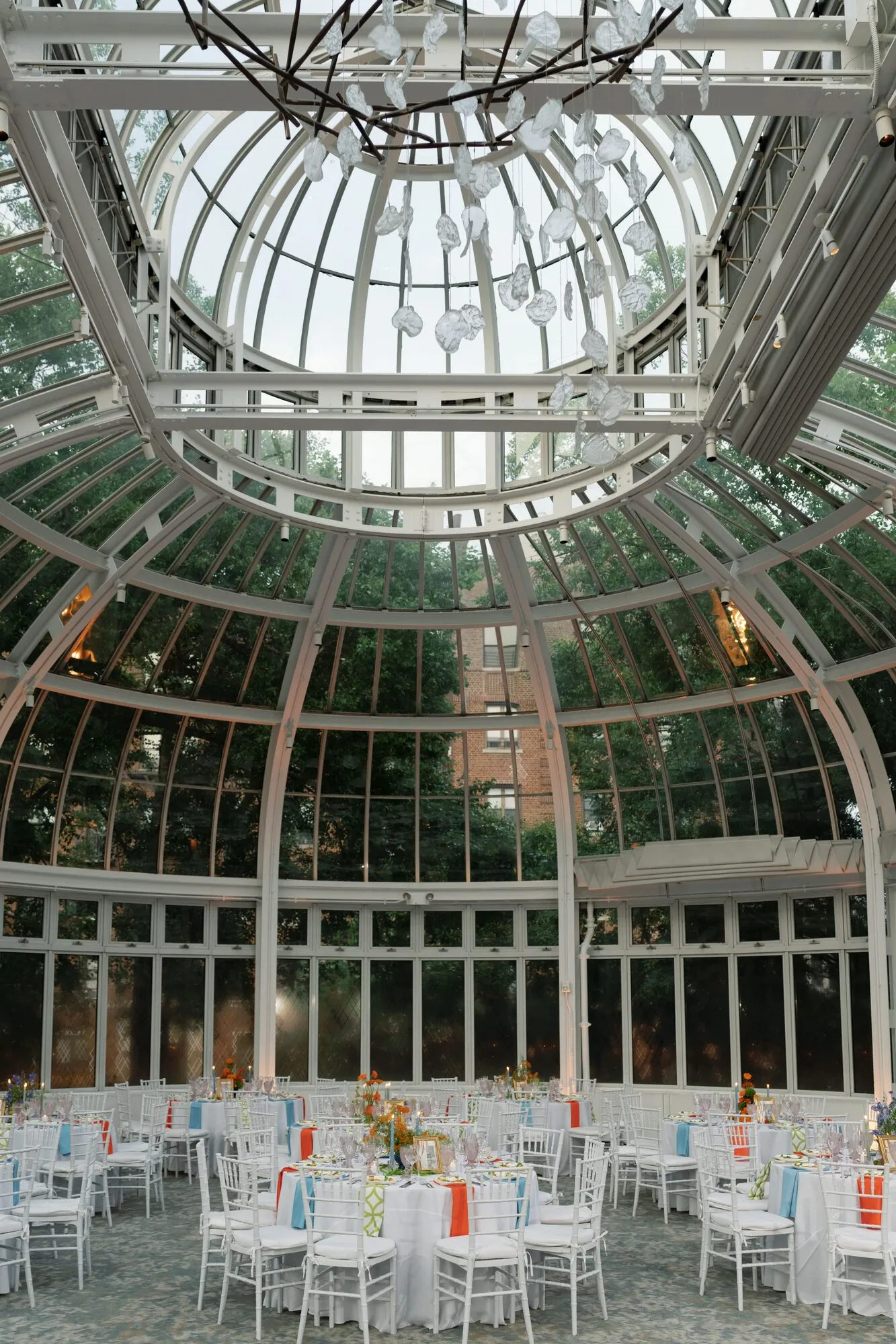 Wide interior view of the Brooklyn Botanic Garden Palm House conservatory, its glass dome overhead framing reception tables dressed in orange, blue and green linens.