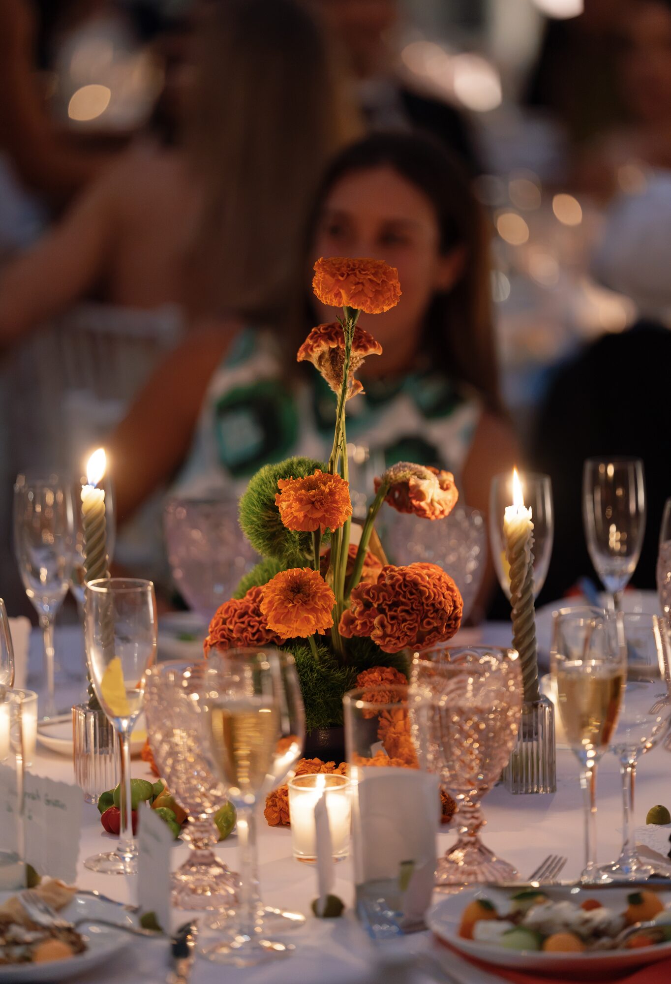 Evening reception table centerpiece of orange zinnias and marigolds surrounded by candlelight, with guests seated in the background.