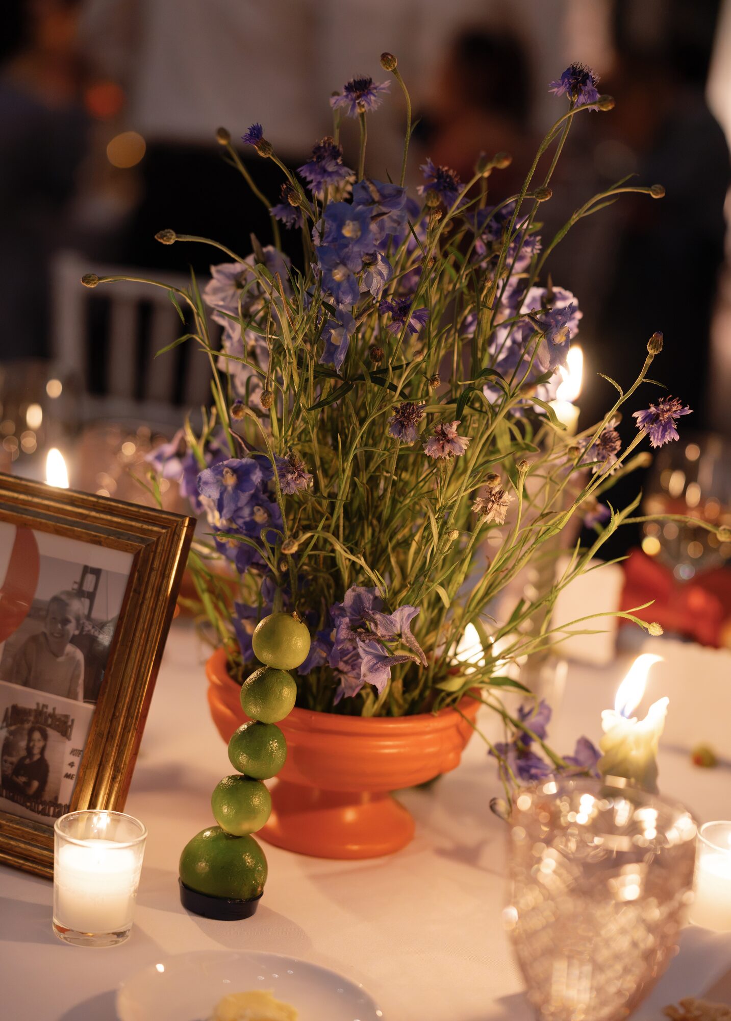 Evening reception centerpiece of purple larkspur and cornflower in a footed orange urn, styled with a stacked lime garnish, votive candles and a vintage photo in a gold frame.