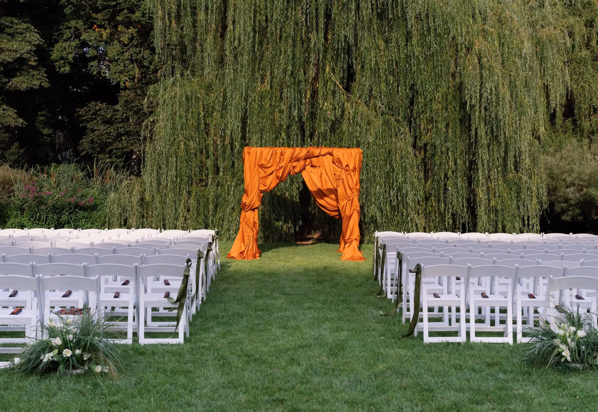 Empty ceremony aisle lined with white folding chairs leading to a chuppah draped in bold orange fabric beneath a weeping willow tree at Brooklyn Botanic Garden.