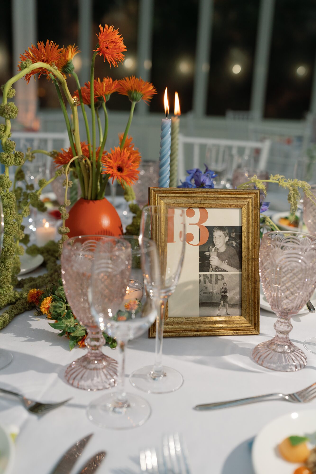 Table number thirteen detail featuring orange gerbera daisies in a moss-wrapped vase, a vintage photo in a gold frame, blue taper candles and blush glassware.