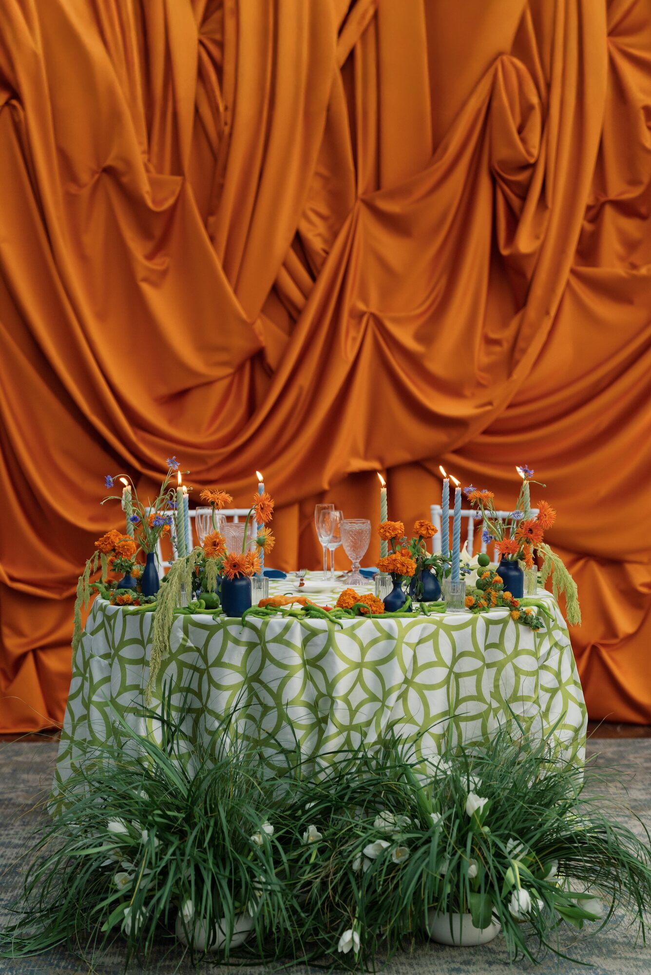 Sweetheart table set against a rippling orange fabric backdrop, styled with a green quatrefoil-print linen, navy bud vases, orange florals and blue taper candles.