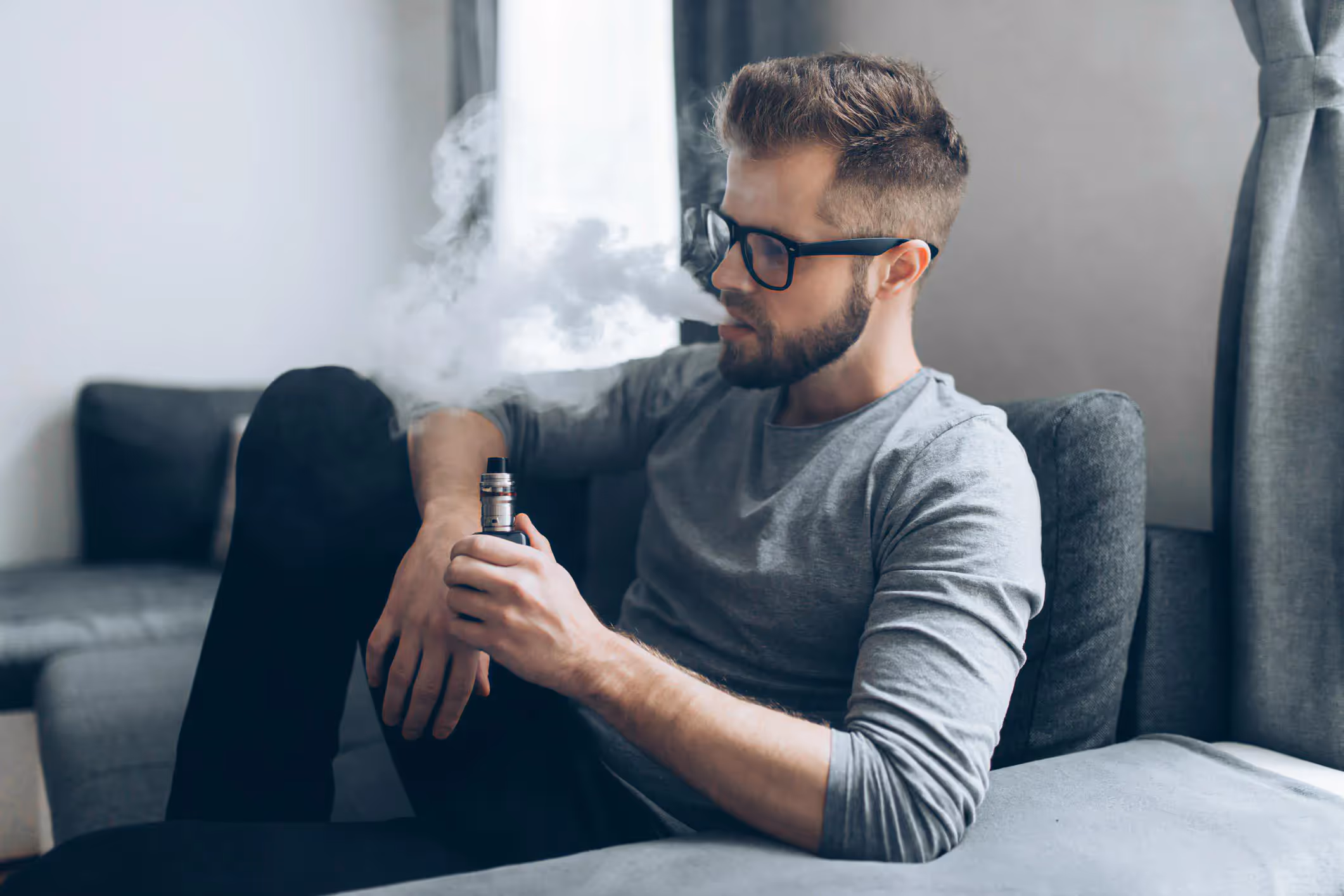 A young man uses a vape device and exhales a cloud of smoke. 