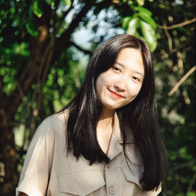 Smiling woman with long black hair wearing a beige shirt standing outdoors with green foliage background.