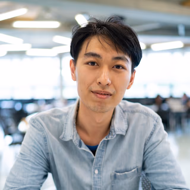 Young man with dark hair wearing a light blue denim shirt looking at the camera in a bright office setting.