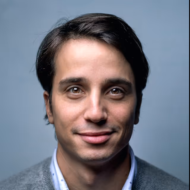 Portrait of a smiling young man with dark hair wearing a grey sweater and light blue collared shirt against a plain blue background.