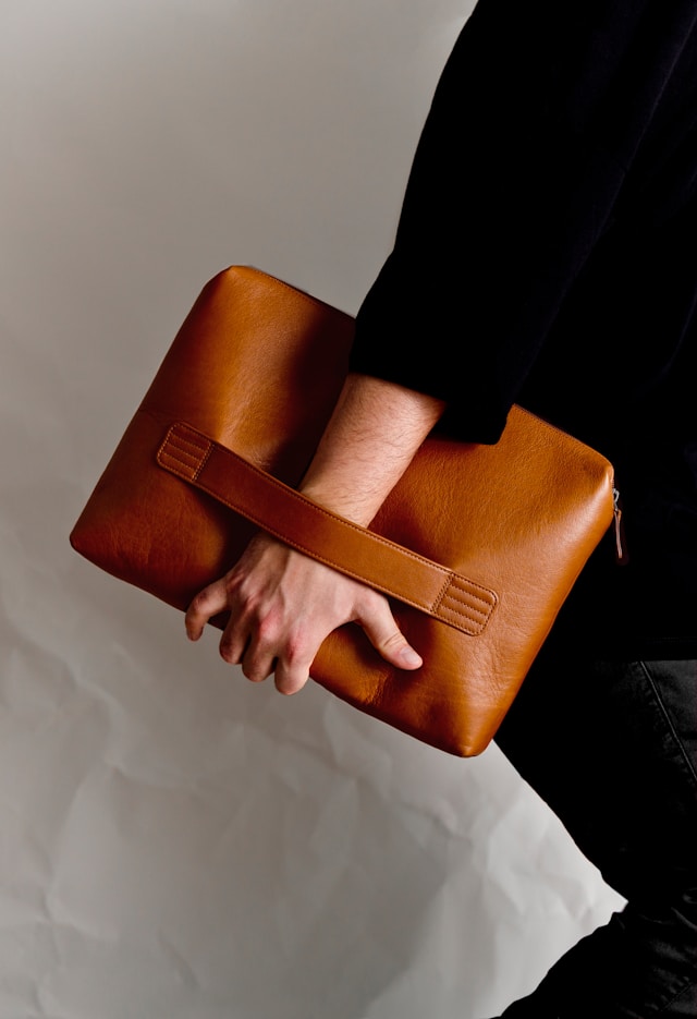 Person wearing a black sleeve holding a tan leather clutch bag with a horizontal strap on a neutral background.