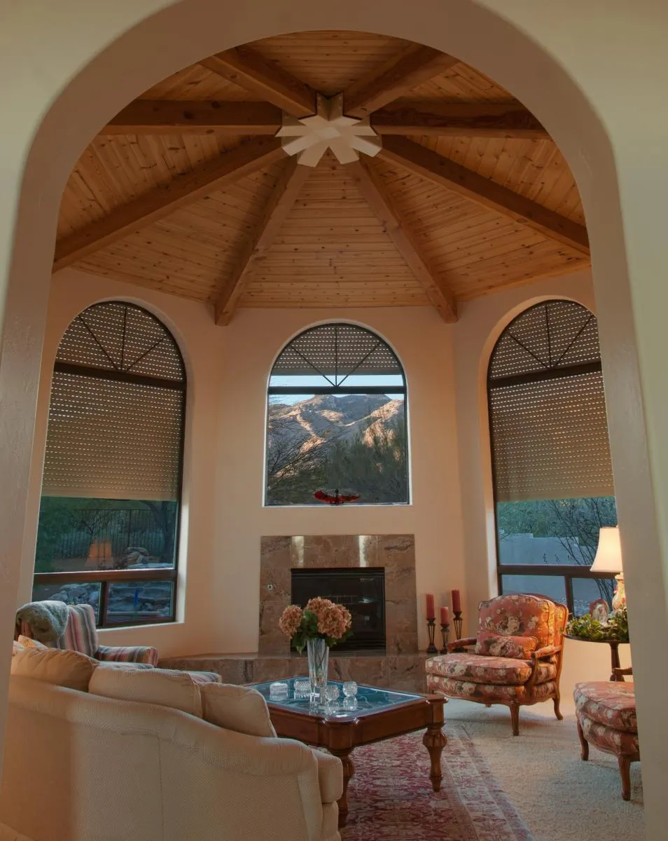 Rustic living room with wood ceiling, three arched windows with rolling shutters, mountain view.