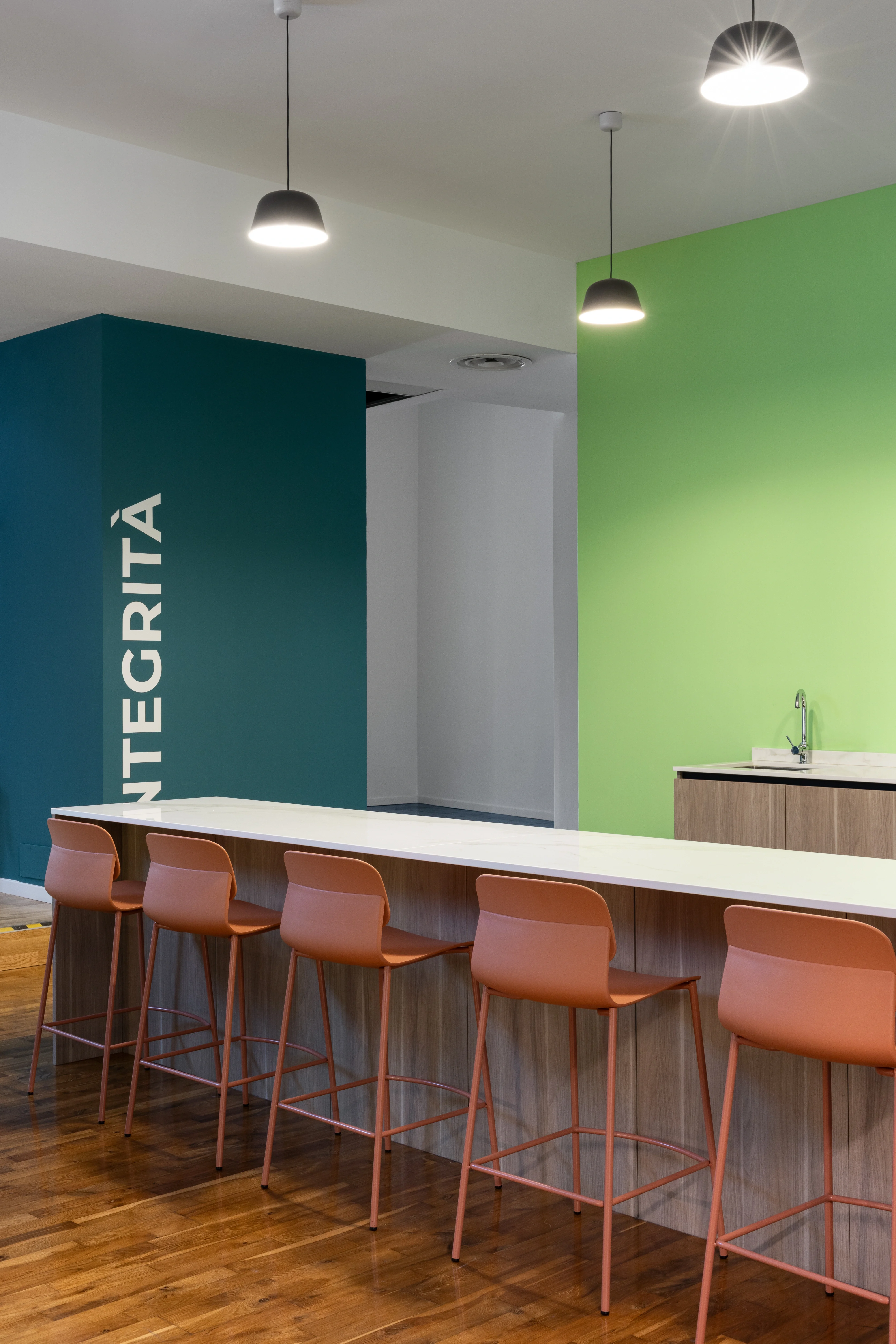 Espace café d’entreprise à Milan avec comptoir en bois clair, chaises hautes terracotta et murs vert et bleu signés Slean pour Ogury.