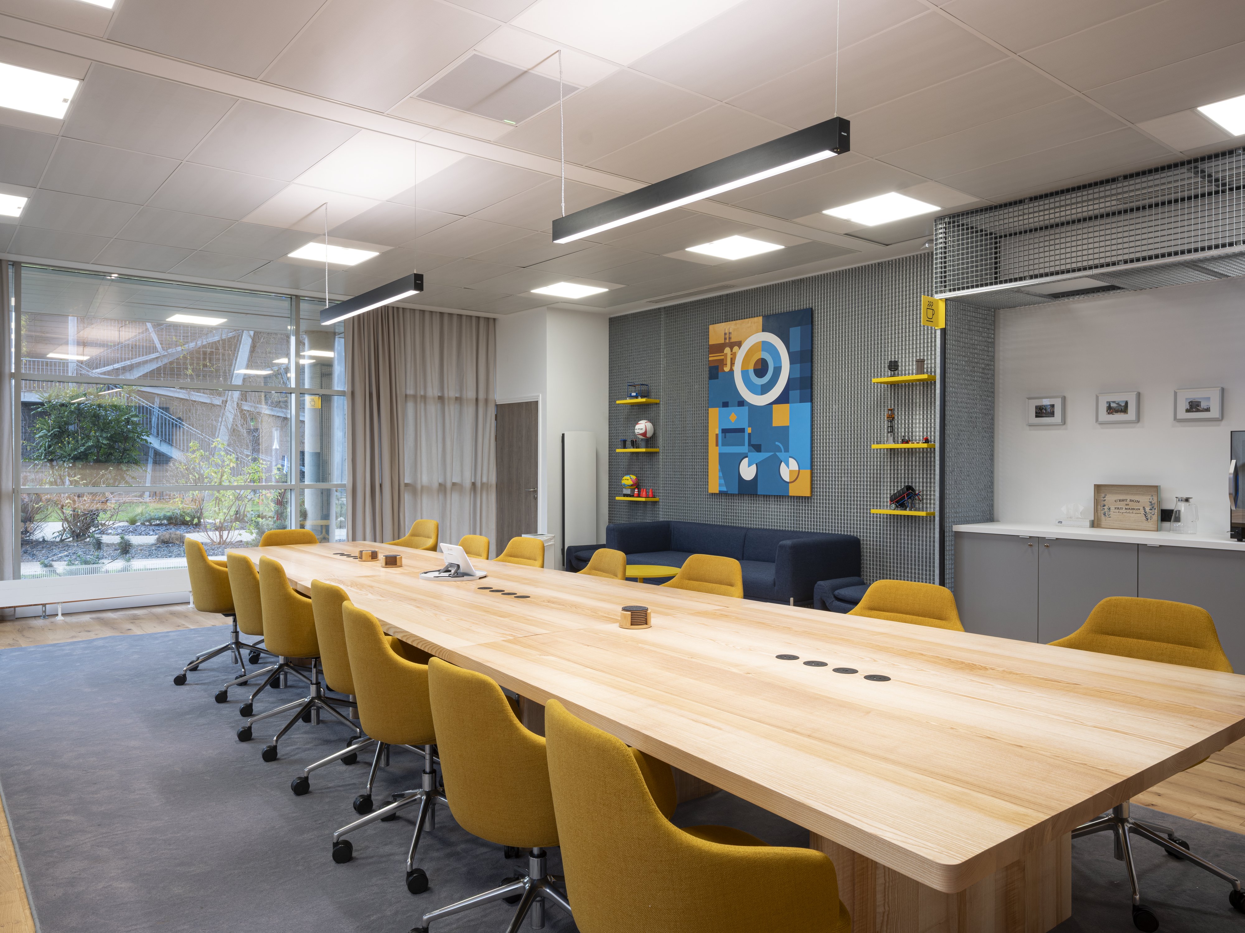 Board room Manutan rénovée : longue table de réunion en bois clair, fauteuils jaunes, éclairage suspendu, coin lounge avec canapé bleu, œuvre murale et grandes baies vitrées.