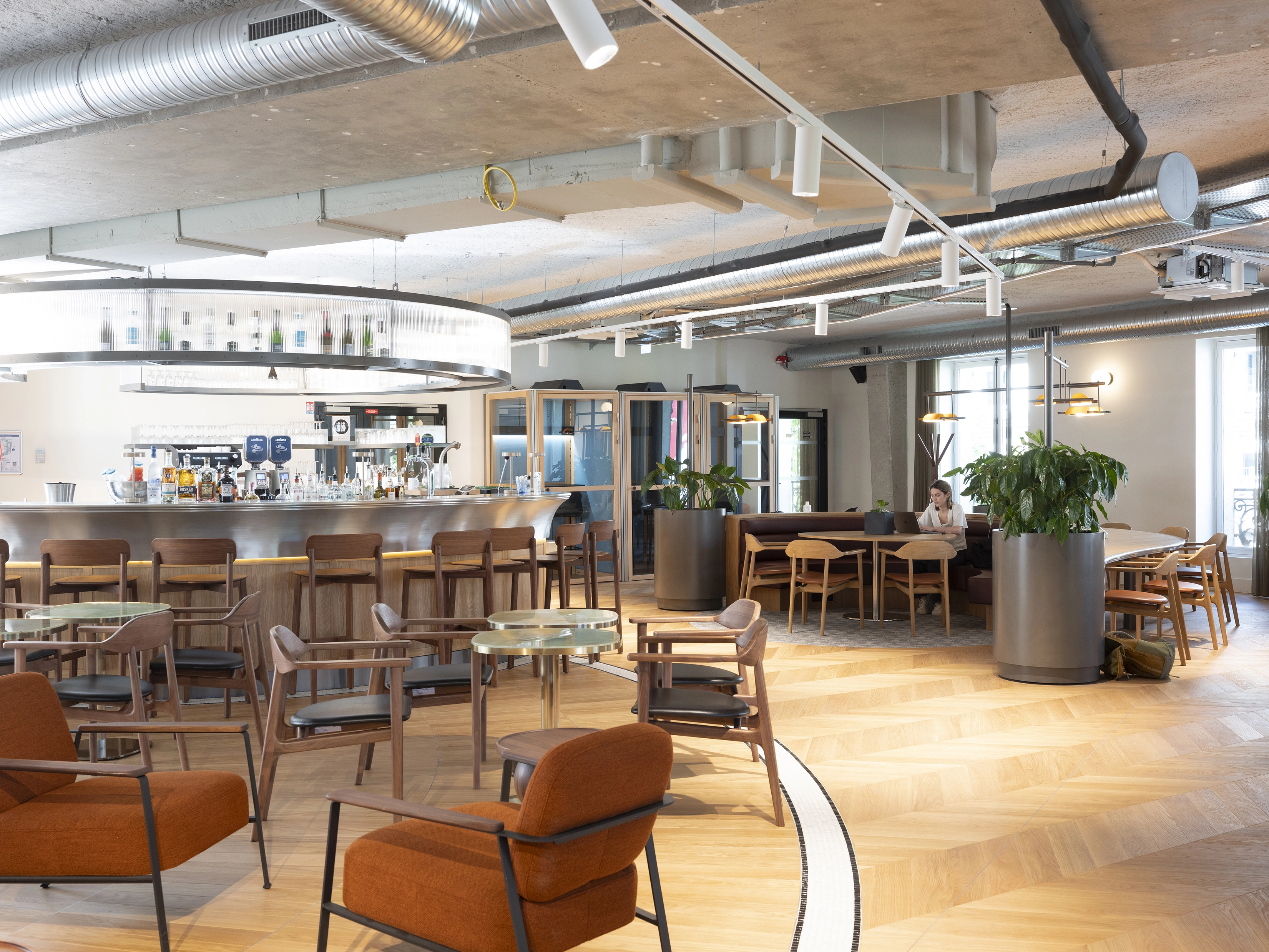 Espace café-lounge chez Motier Ventures : bar central avec tabourets, tables et fauteuils, grandes plantes, parquet en chevrons et plafond industriel ; au fond, modules vitrés type cabines/box (installation Métropole)