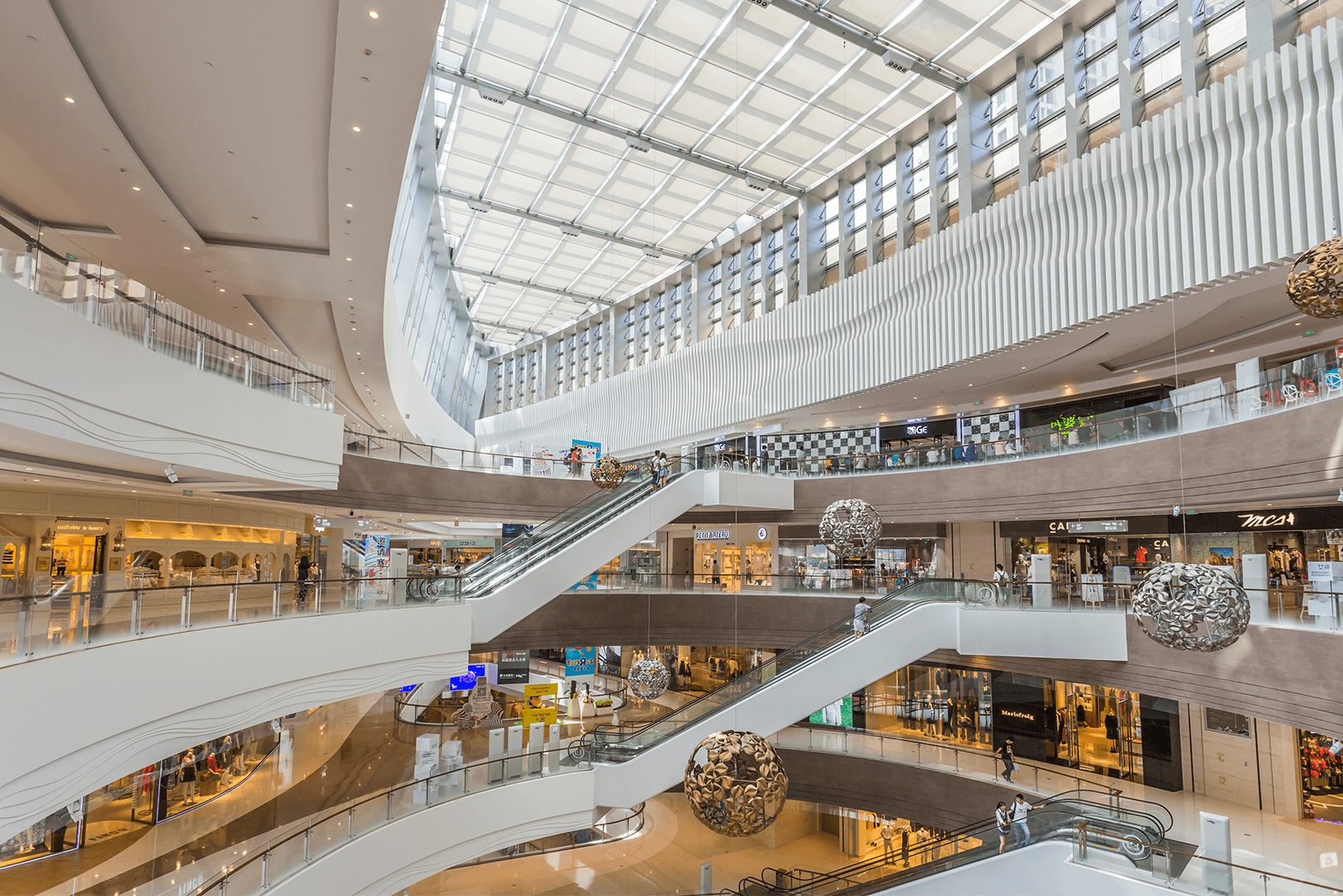 Interior de shopping center moderno com múltiplos andares, escadas rolantes e lojas ao fundo, representando o ambiente de varejo em pontos comerciais de alto custo de ocupação
