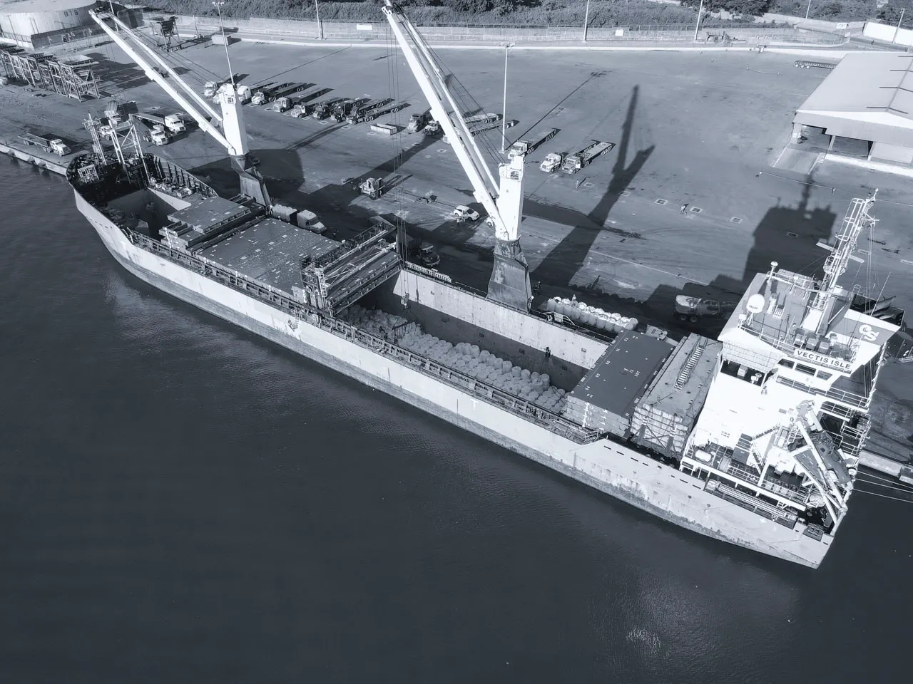 Aerial view of the Vectis Isle vessel during port operations: Ship cranes loading breakbulk cargo units into the hold for international transit.
