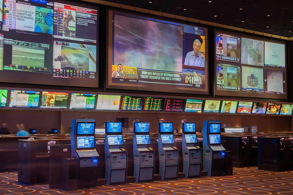 Aliante sportsbook before the remodel &mdash; legacy projector-based display wall with multiple screens and a row of betting kiosks across patterned carpet