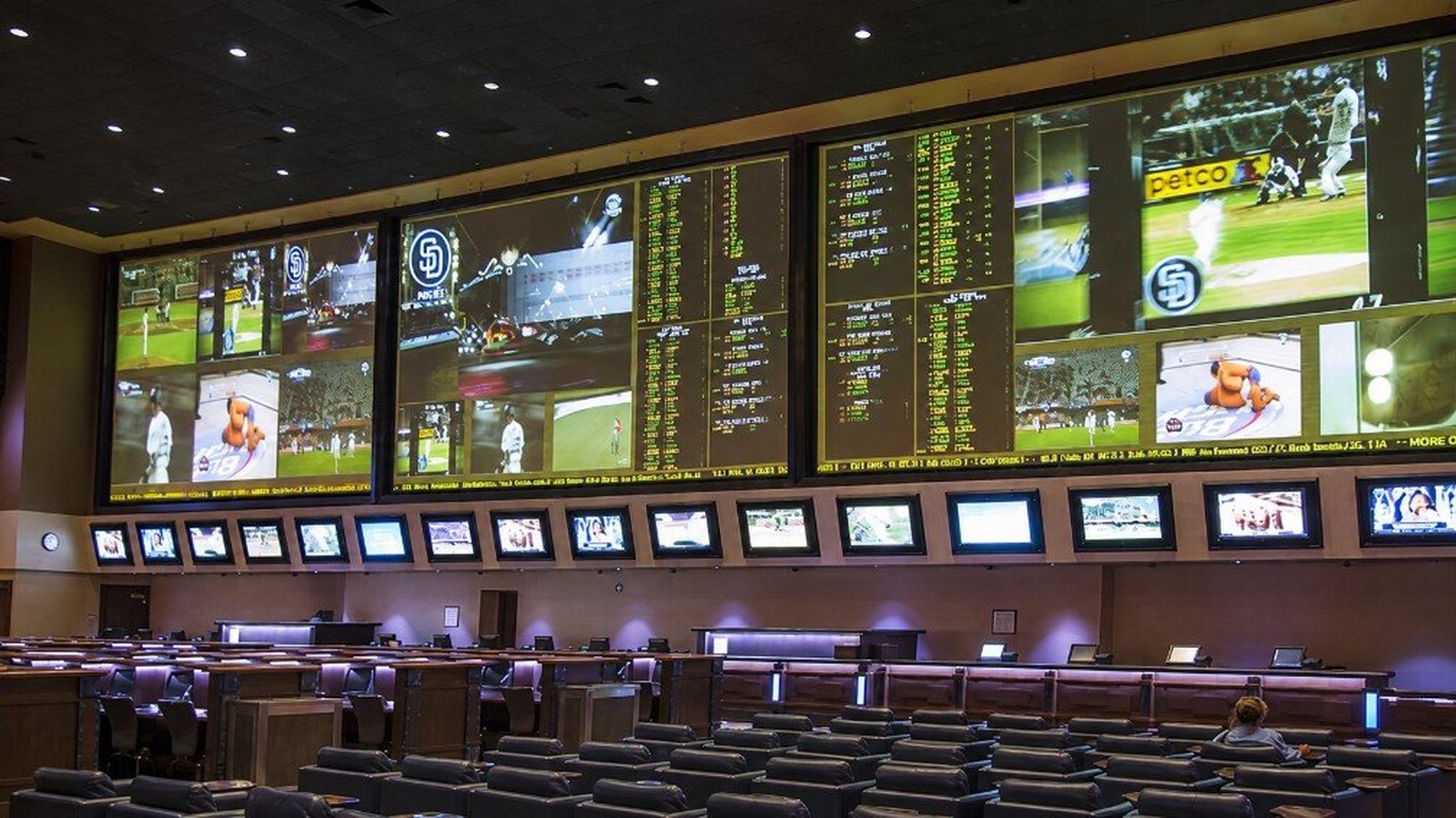 Aliante sportsbook before the remodel &mdash; curved wall of multiple projection screens with a long row of small monitors forming the odds ticker, theater-style seating in the foreground