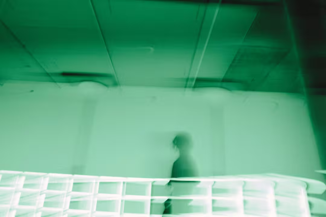 Blurred silhouette of a person walking behind a white fence in a green-tinted room interior.