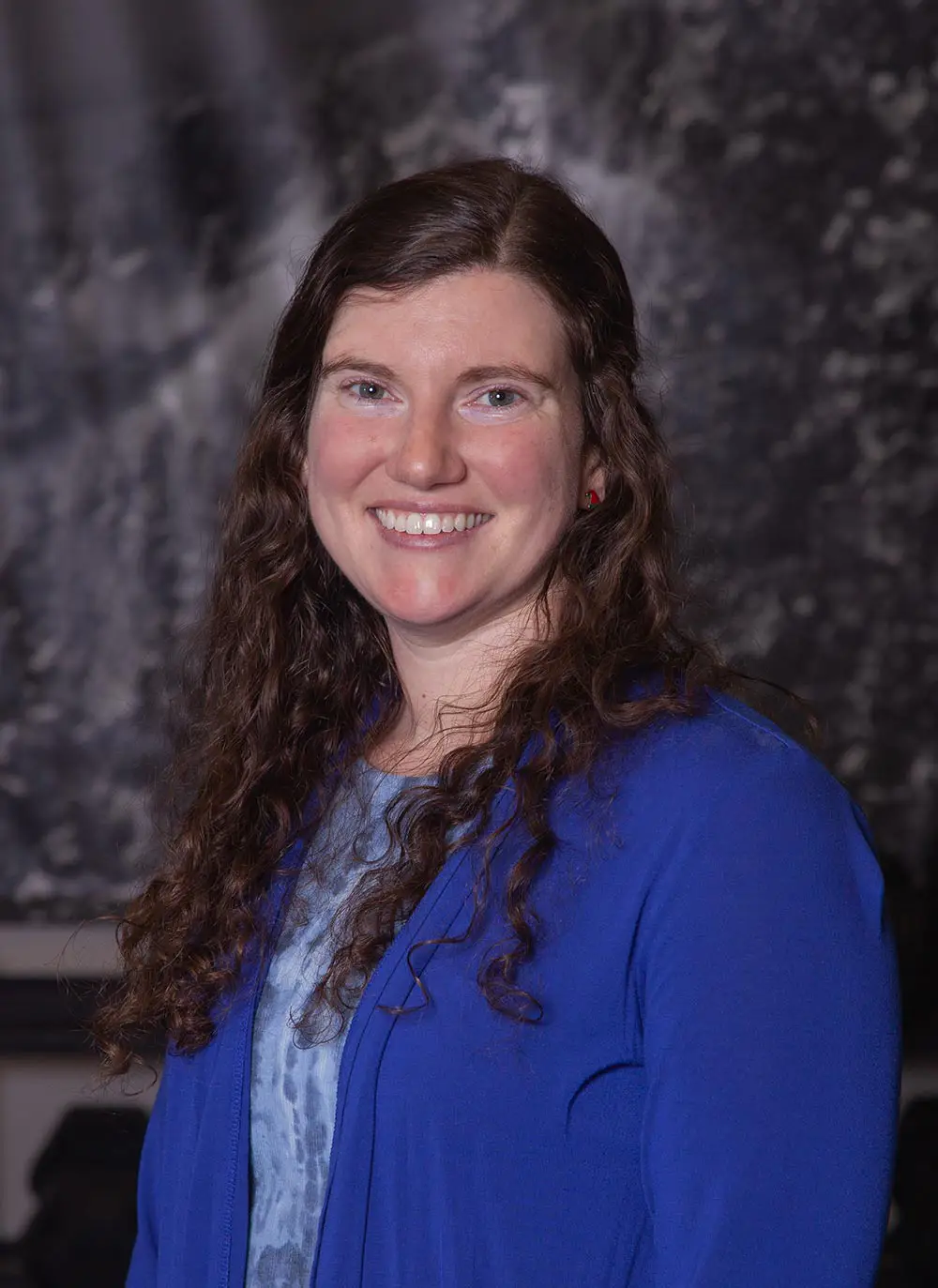 Smiling woman with long curly brown hair wearing a blue cardigan over a patterned top against a dark blurred background.