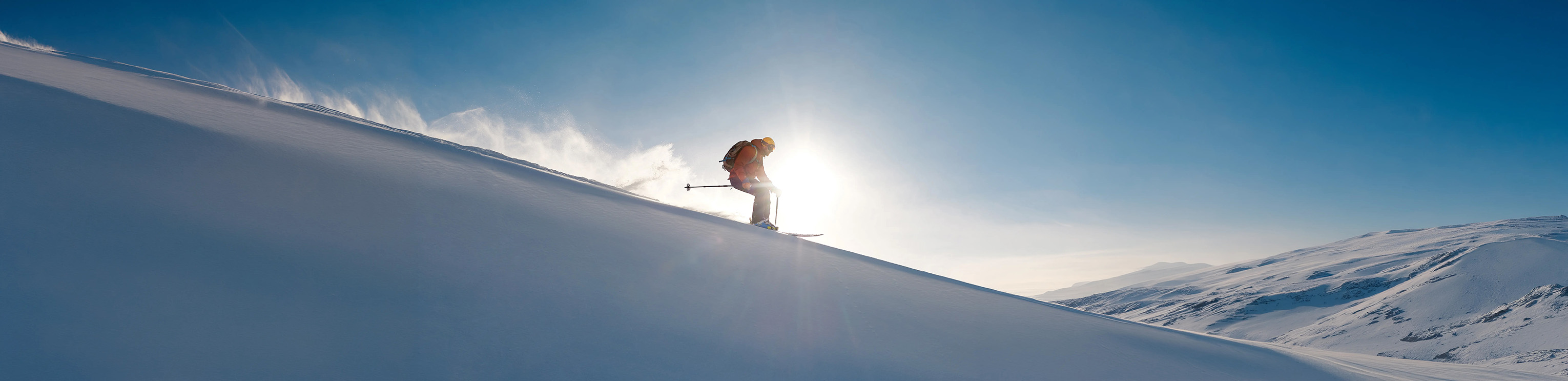 Skier descending a snowy mountain slope with the sun shining directly behind them.