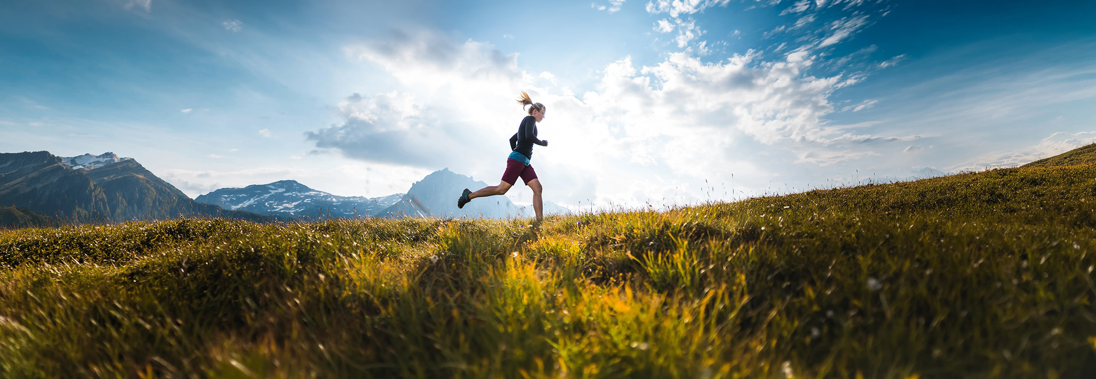 Woman trail running on a grassy hill with mountains and a blue sky with clouds in the background.