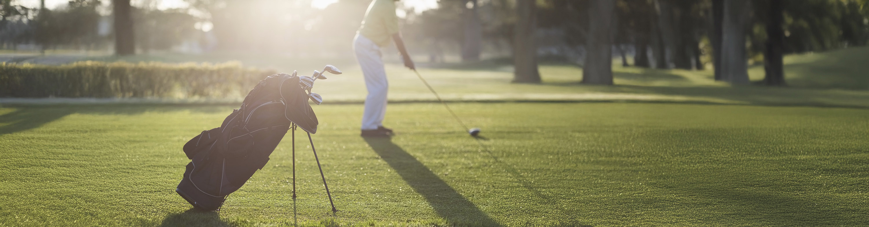 Golf bag with clubs on grass in foreground, golfer preparing to putt in the background during sunny day.