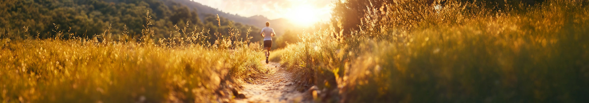 Person jogging on a narrow dirt path through golden grass at sunset with hills in the background.