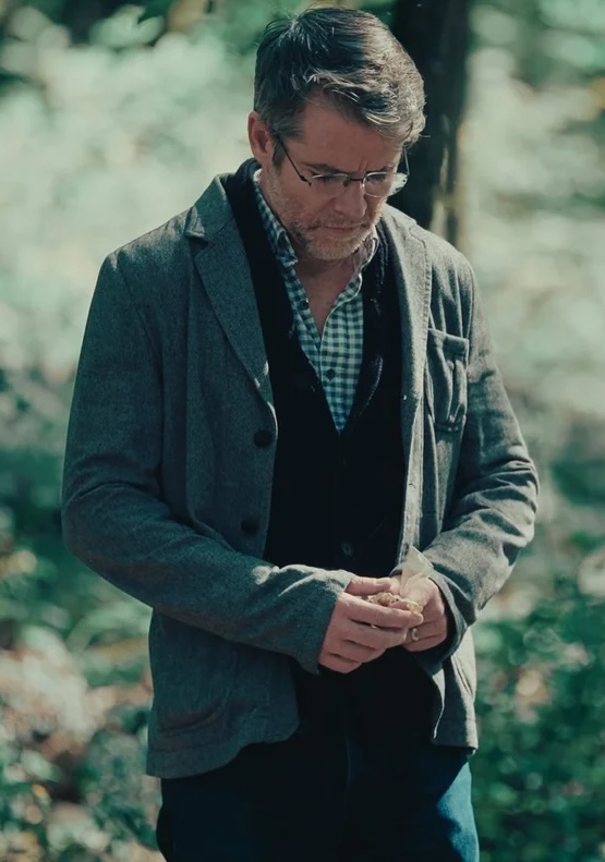 Man with glasses wearing a gray jacket and checkered shirt looking down while holding an object outdoors.