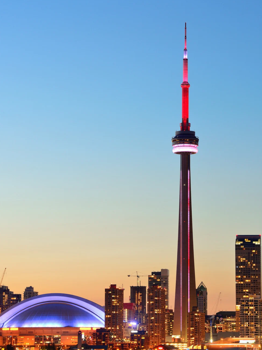 Toronto skyline at dusk featuring the illuminated CN Tower with red and pink lights and nearby buildings with glowing windows, against a clear gradient sky from orange near the horizon to blue above.