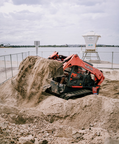 Red Kubota skid-steer loader moving large pile of sand near a fenced waterfront with lifeguard tower.