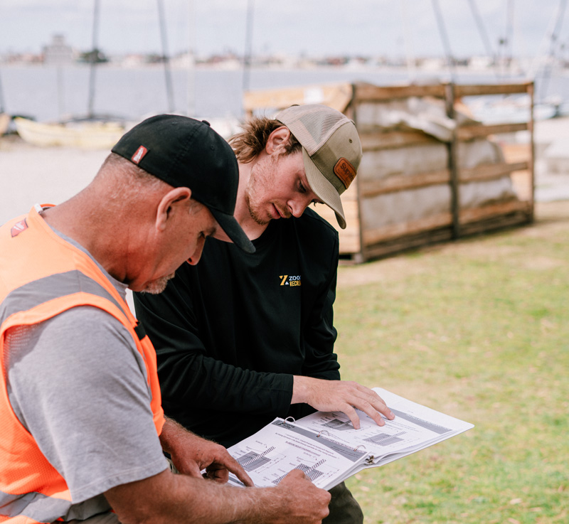 Two men outdoors reviewing architectural plans on a clipboard near a body of water.