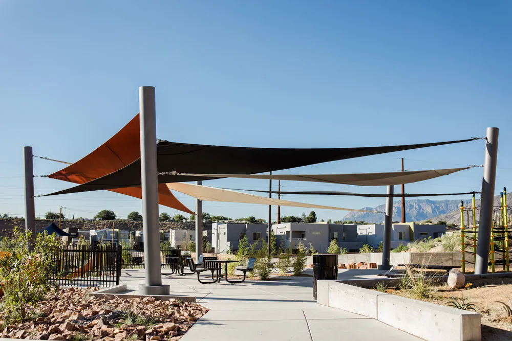 Outdoor seating area with multiple triangular shade sails in black, brown, and beige stretched between tall metal poles under clear blue sky.