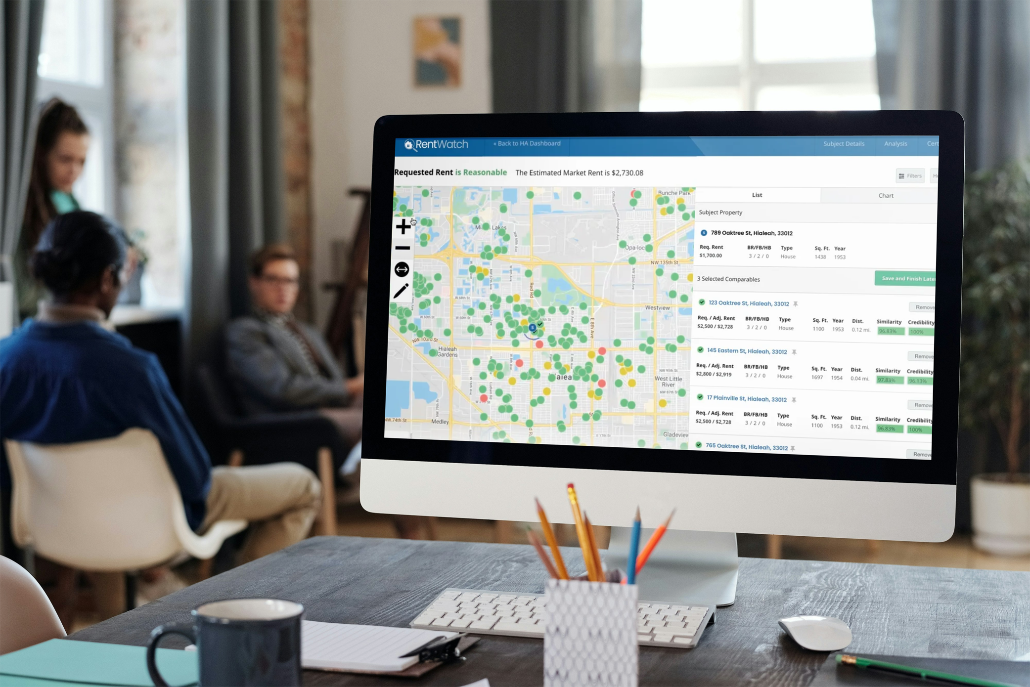 image of an office computer with RentWatch on the screen, a meeting with employees are having a meeting in the background.