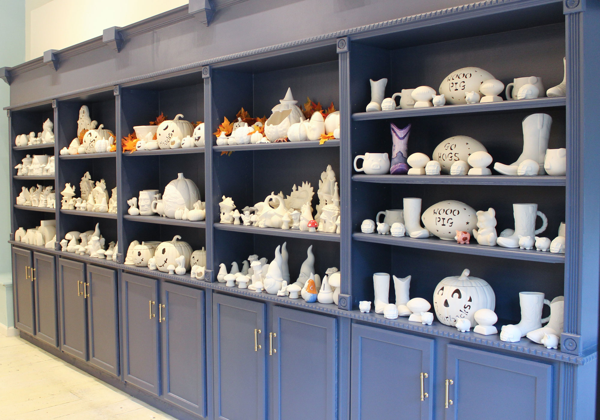 Blue shelving unit filled with unpainted ceramic decorations including pumpkins, boots, footballs, mugs, pigs, and seasonal fall items.