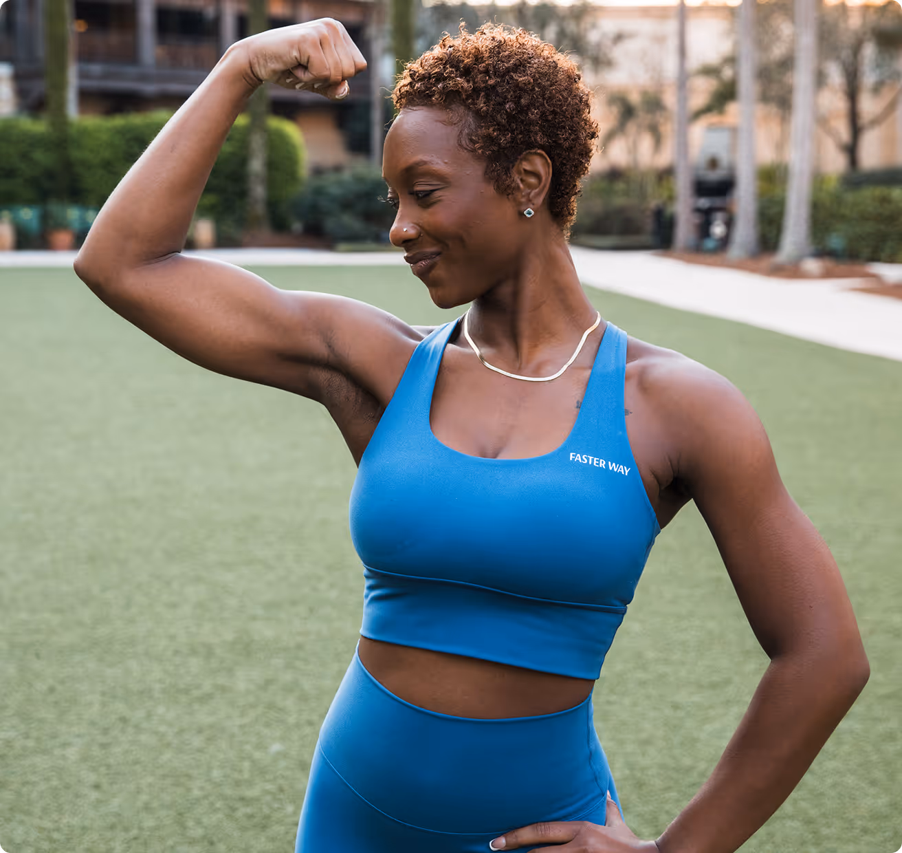 Smiling woman in blue workout clothes flexing her arm muscle outdoors on grass.