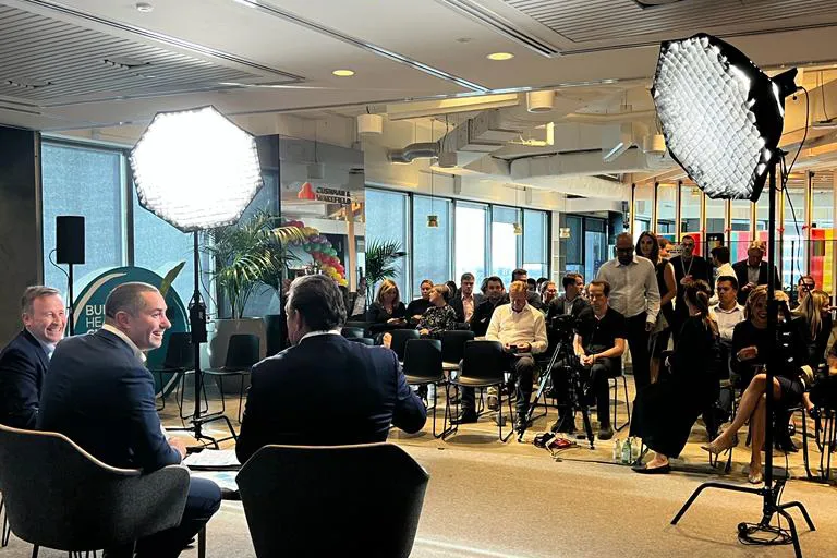 Three men in suits seated in front of an audience in a well-lit modern room with professional lighting equipment set up for a panel discussion or interview.