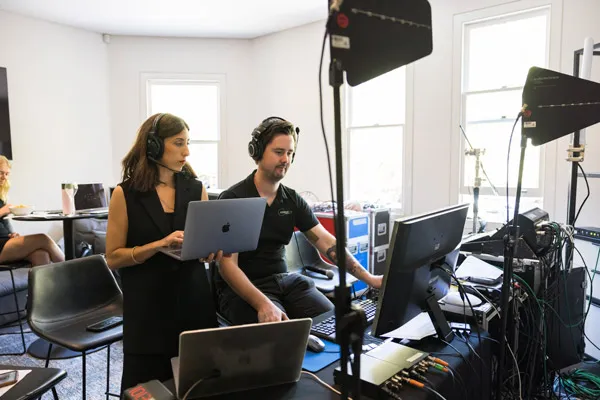Two people wearing headsets working on laptops and a desktop computer in a bright room with professional lighting equipment.