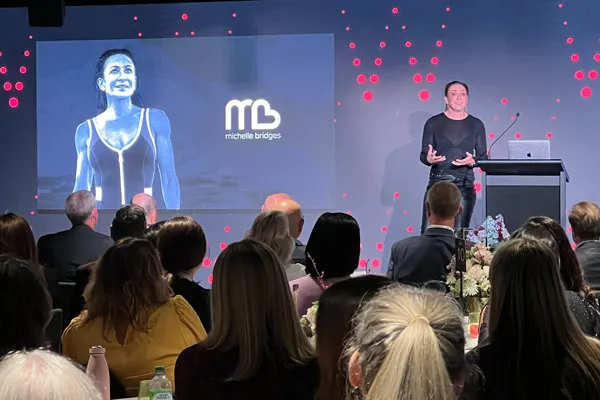 A woman speaker presenting on stage to an audience with a projected image of a smiling Michelle Bridges in athletic wear and her logo.