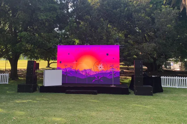 Outdoor stage with large colorful LED screen displaying a sunset graphic with palm trees, surrounded by speakers and trees in a park.