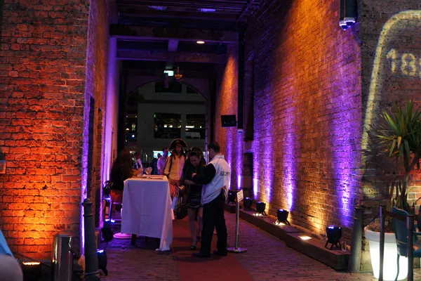 People gathered in a narrow brick corridor lit with orange and purple uplights, with security personnel checking guests at a table.