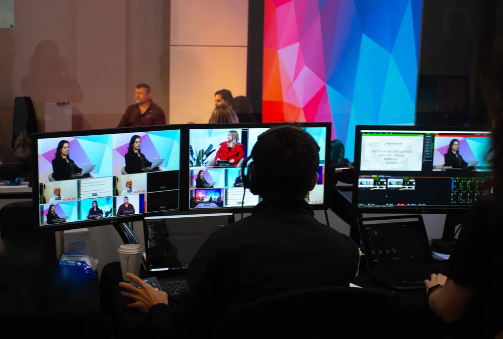 Person wearing headphones editing a live broadcast on three monitors showing multiple video feeds and participants.