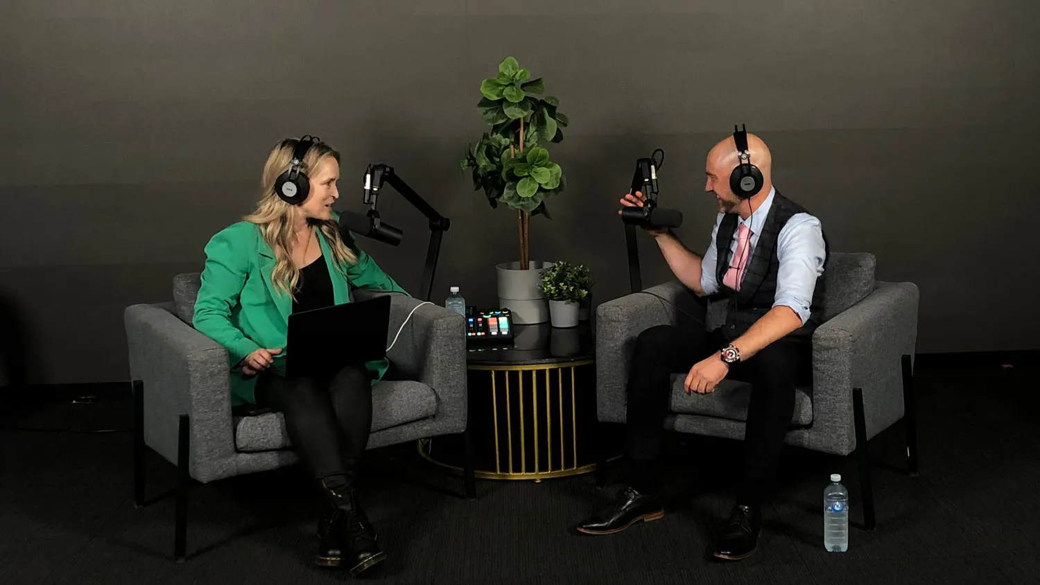 Two people wearing headphones and speaking into microphones while seated in armchairs with a small table and plants between them in a podcast recording studio.