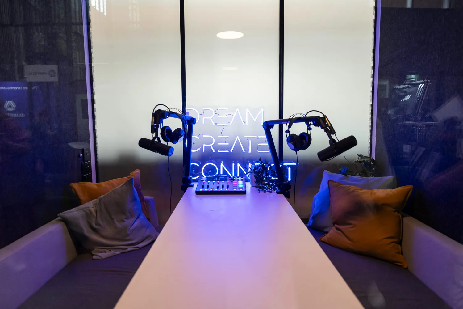 Podcast recording setup with two microphones, headphones, and colorful pillows on couches beside a table with a neon sign reading 'Dream Create Connect.'