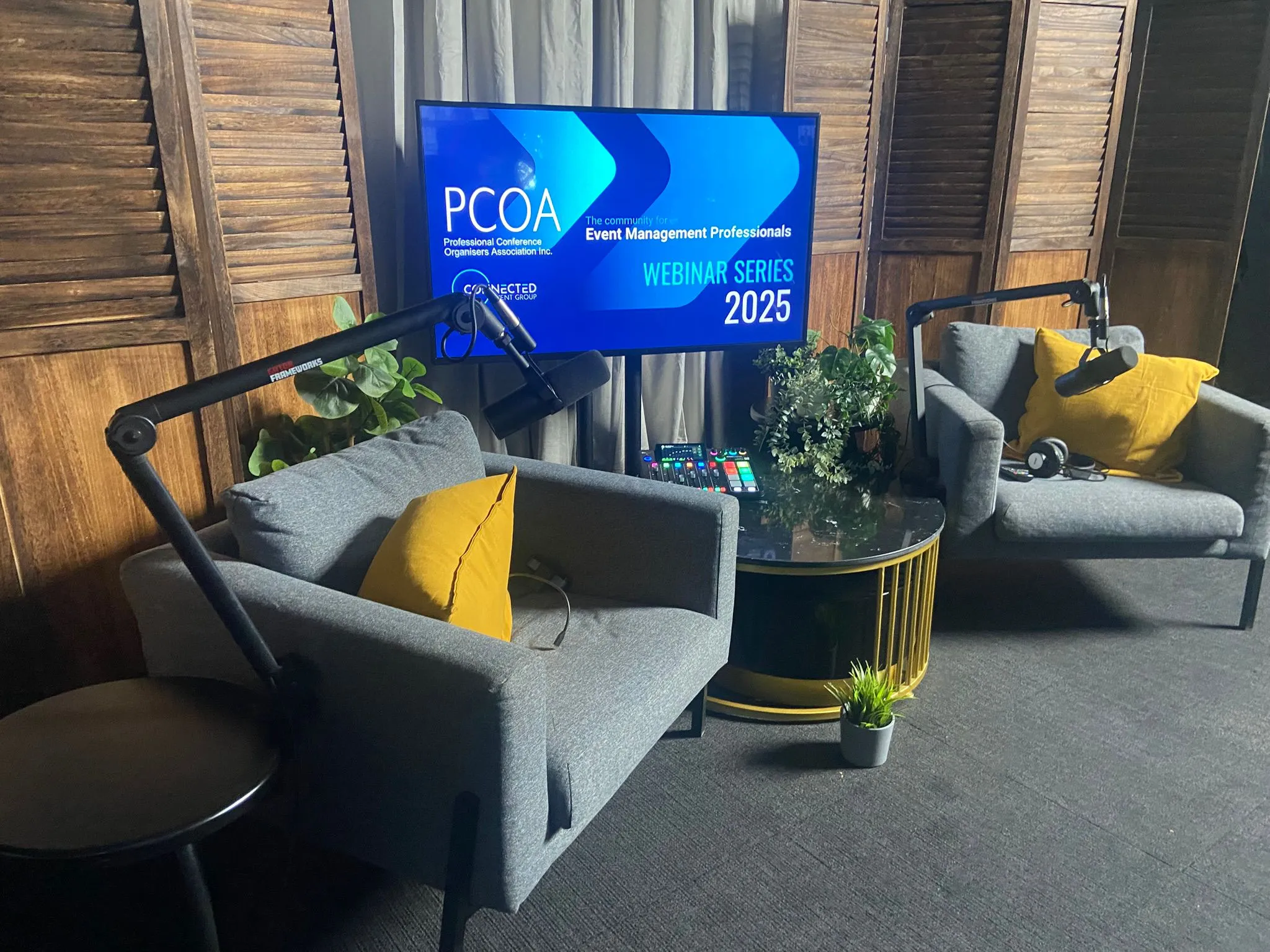 Podcast studio setup with two gray armchairs with yellow cushions, black microphones on adjustable arms, a round glass coffee table with plants, and a screen displaying PCOA Webinar Series 2025.