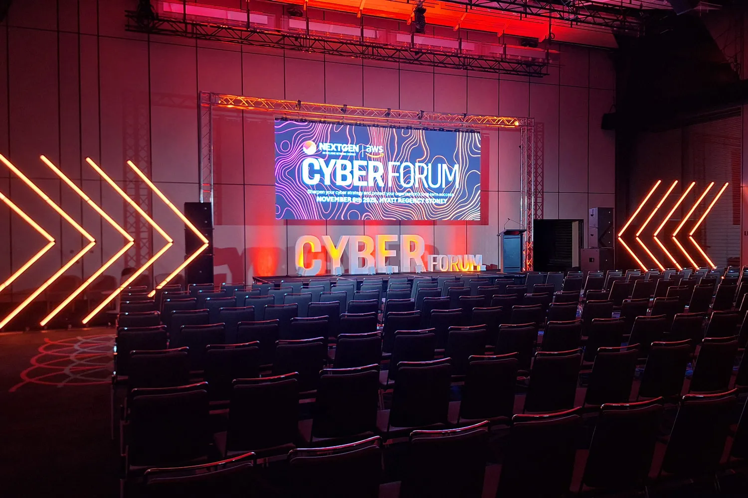 Empty conference room with chairs facing a stage lit with orange lights and a Cyber Forum screen.