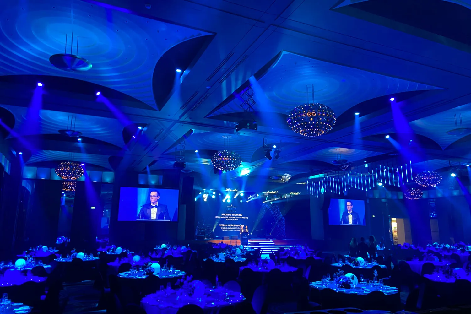 Blue-lit banquet hall with round tables and a man speaking on stage shown on large screens.