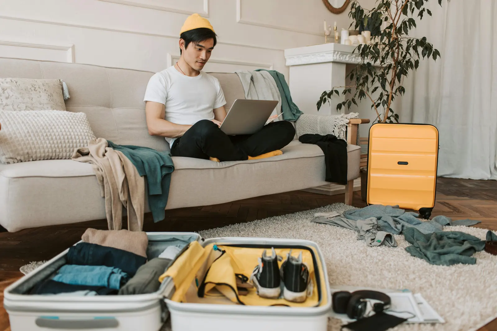 Picture of an Asian man sitting on the sofa using the laptop after he returned from the holiday. The unpack bags are still on the floor while he is hyperfocusing on his tasks.