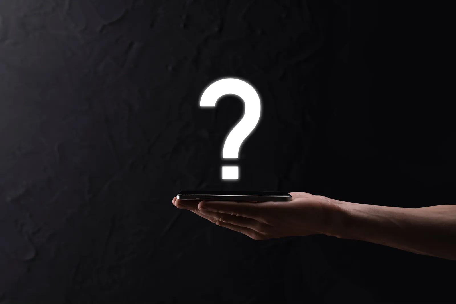Picture of a hand on a dark background holding a mobile phone and above having a question mark. Picture for the FAQ section of this article.