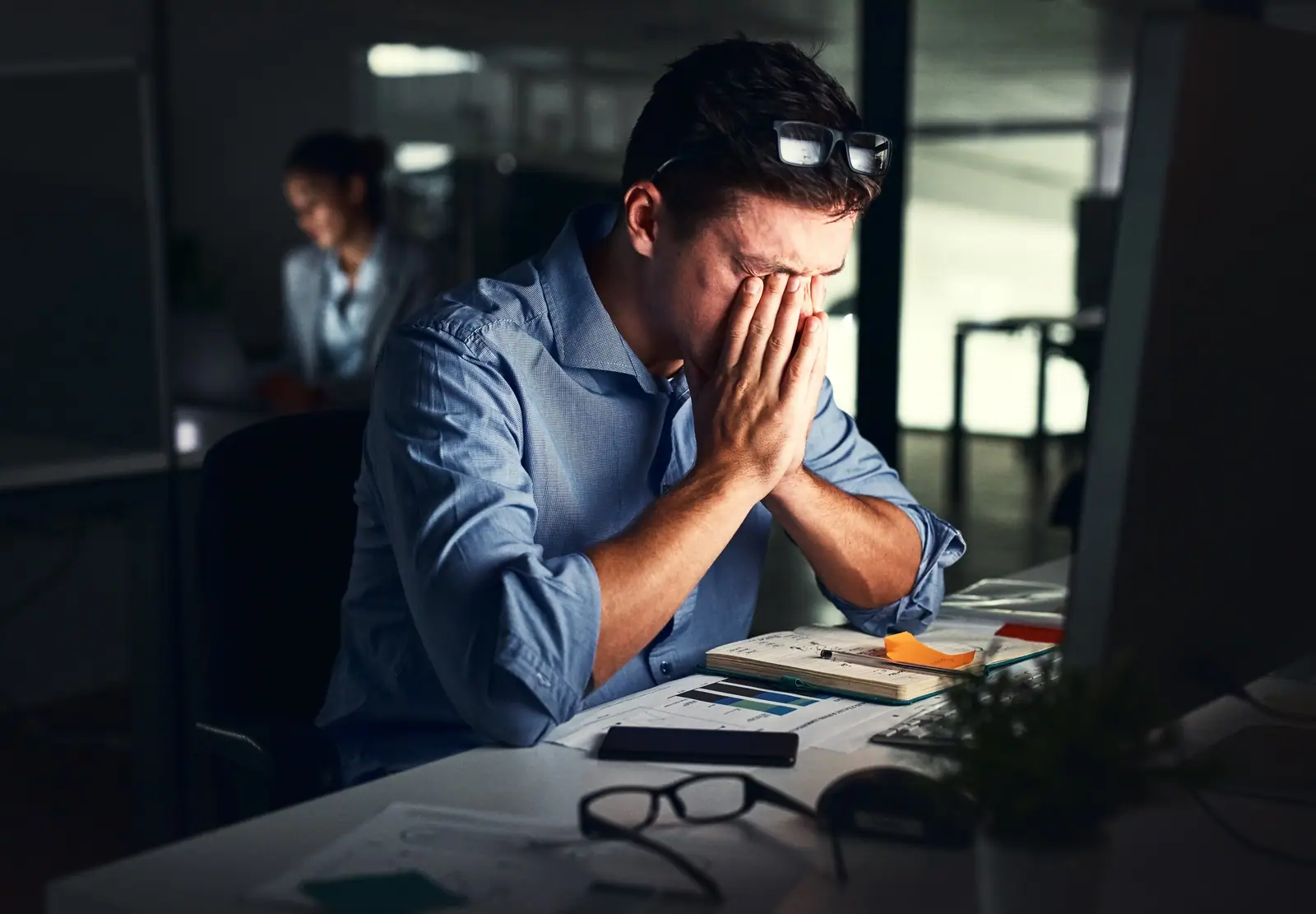 Professional male, and in a background a professional female staying late at night at risk of developing burnout.