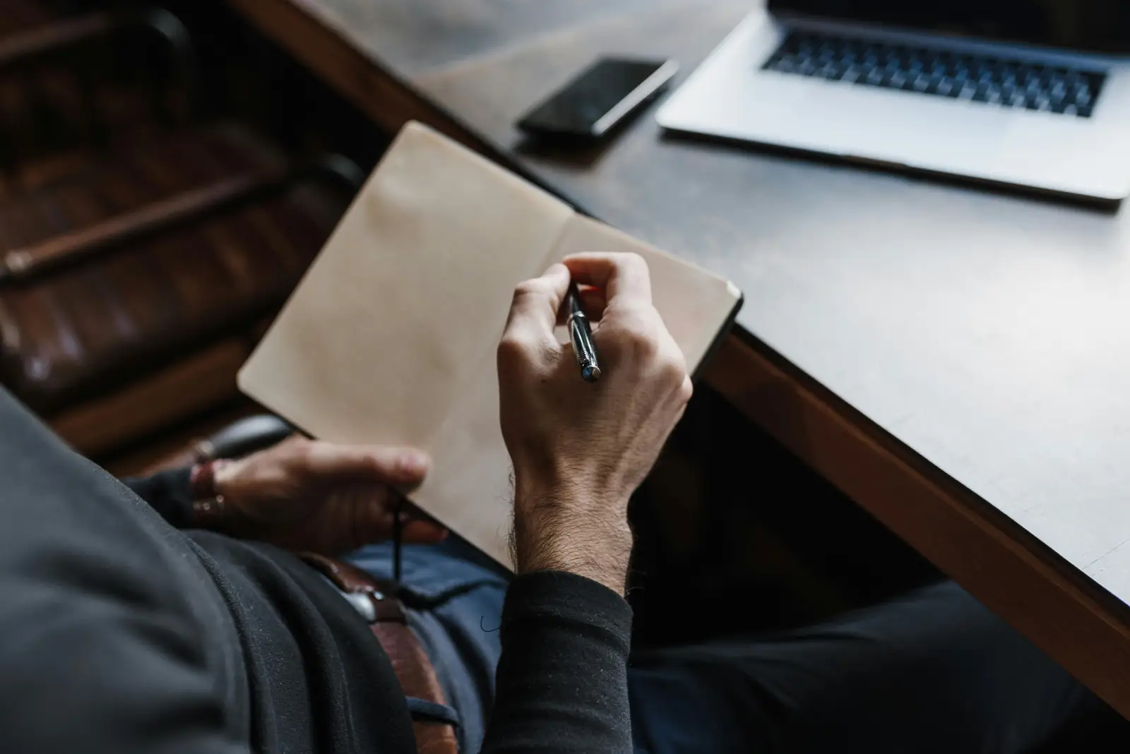 Picture of a man taking notes with pen and paper while also having on his desk a laptop.
