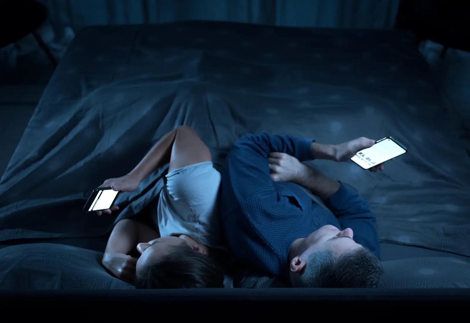 picture of a couple sitting in bed at night disconnected from each other while suing the mobile phones. 