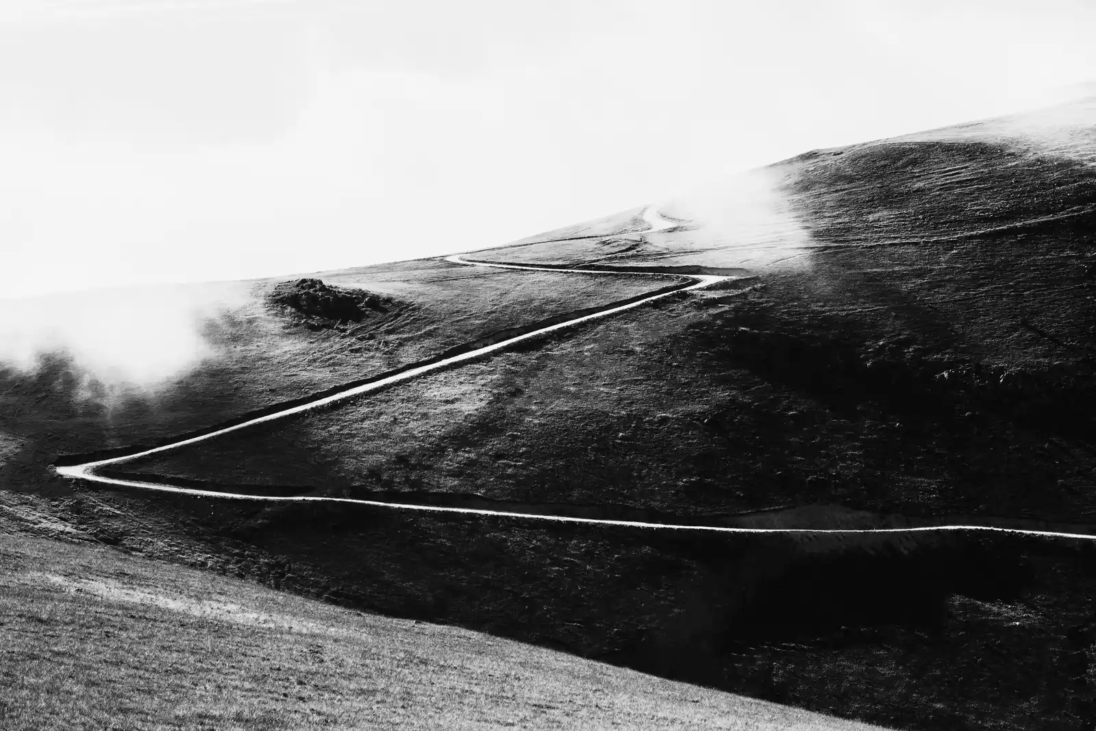 Monochrome landscape of a winding mountain dirt road ascending through mist toward an open horizon, representing non-linear psychological resilience.